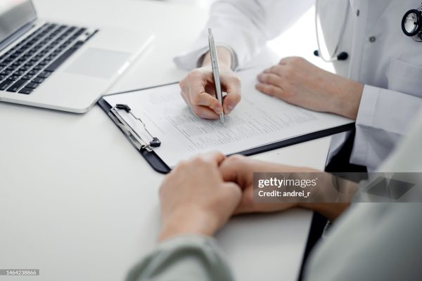Doctor and patient sitting at the desk in clinic office. The focus is on female physician's hands filling up the medication history record form, close up. Medicine concept.